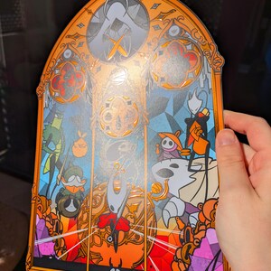 May include: A stained-glass style art print featuring characters from the video game Hollow Knight. The artwork has an orange border and a blue and orange background. The print includes the text "Catterine Art".