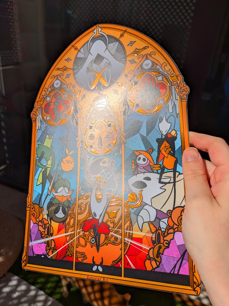 May include: A stained-glass style art print featuring characters from the video game Hollow Knight. The artwork has an orange border and a blue and orange background. The print includes the text "Catterine Art".