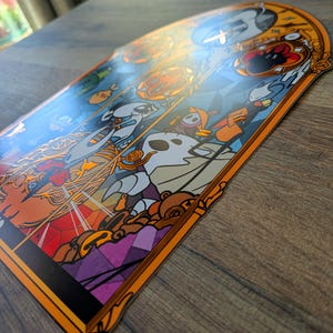 May include: A decorative sticker with a stained-glass design featuring cartoon characters in shades of orange, white, blue, and purple. The sticker has an orange border and is placed on a wooden surface.