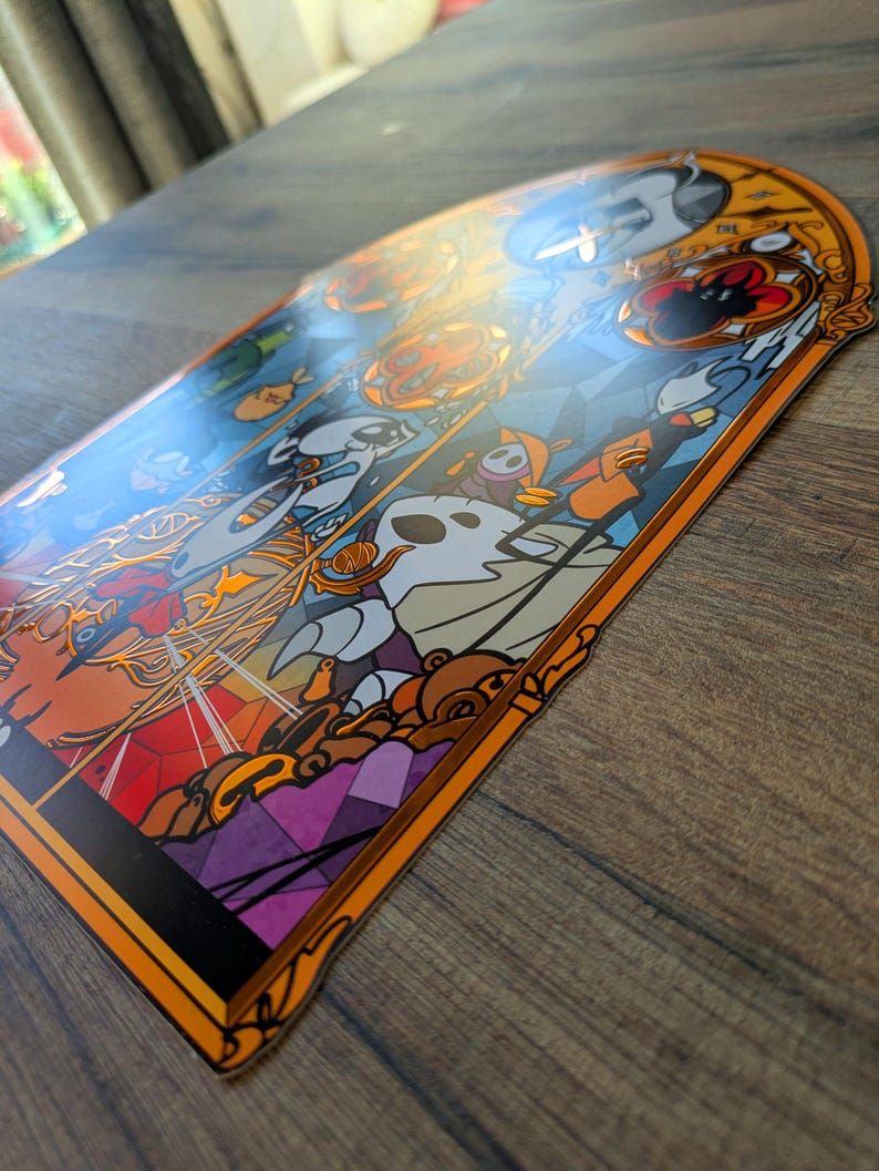 May include: A decorative sticker with a stained-glass design featuring cartoon characters in shades of orange, white, blue, and purple. The sticker has an orange border and is placed on a wooden surface.