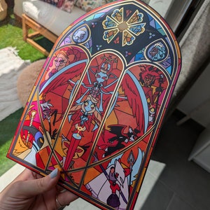May include: A stained glass style print featuring characters from the animated series "Helluva Boss". The print is framed in gold and features a red, orange, and yellow color scheme.
