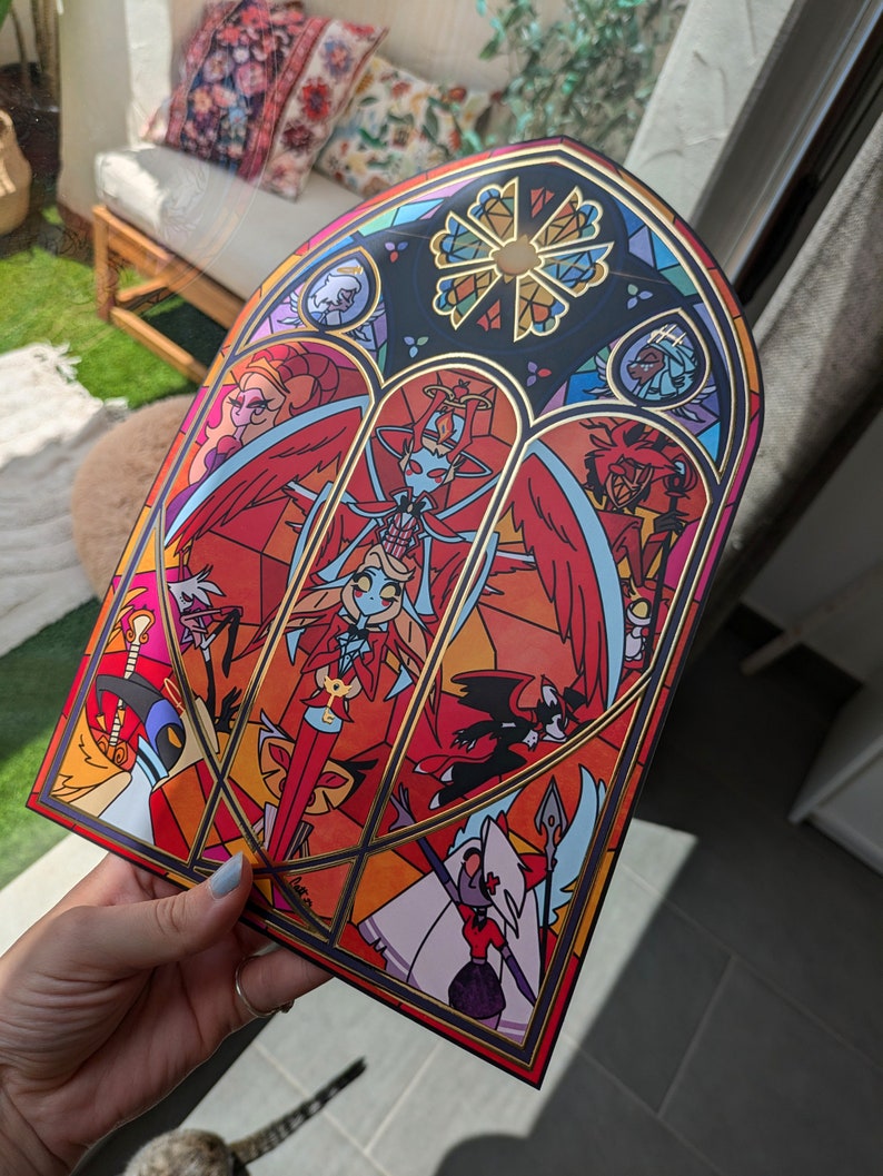 May include: A stained glass style print featuring characters from the animated series "Helluva Boss". The print is framed in gold and features a red, orange, and yellow color scheme.