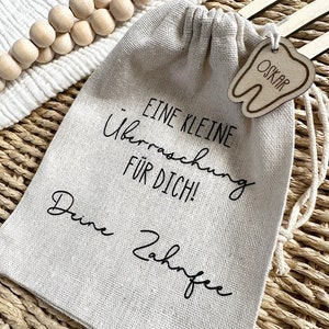 May include: A small, beige linen drawstring bag with a wooden tooth-shaped tag that says "Oskar" hanging from it. The bag has black text that reads "Eine kleine &Uuml;berraschung f&uuml;r dich! Deine Zahnfee".