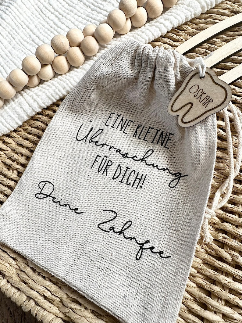May include: A small, beige linen drawstring bag with a wooden tooth-shaped tag that says "Oskar" hanging from it. The bag has black text that reads "Eine kleine &Uuml;berraschung f&uuml;r dich! Deine Zahnfee".