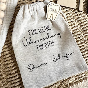 May include: A small white cloth bag with a wooden tooth-shaped tag that says "OSKAR" hanging from it. The bag has black text that reads "Eine kleine &Uuml;berraschung f&uuml;r dich! Deine Zahnfee".