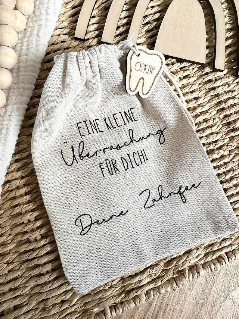 May include: A small white cloth bag with a wooden tooth-shaped tag that says "OSKAR" hanging from it. The bag has black text that reads "Eine kleine &Uuml;berraschung f&uuml;r dich! Deine Zahnfee".