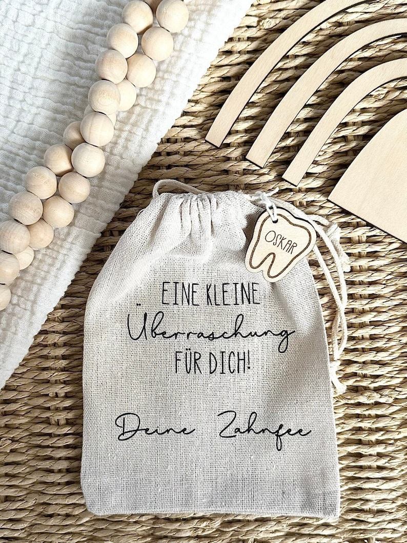 May include: A small white burlap bag with a drawstring closure. The bag has black text that reads "Eine kleine &Uuml;berraschung f&uuml;r dich! Deine Zahnfee". A small wooden tooth with the name "Oskar" is attached to the bag.