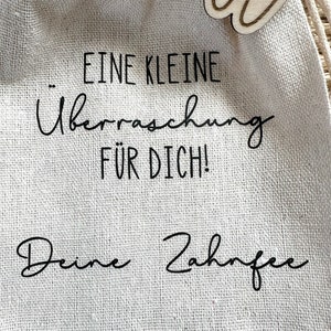May include: A small burlap bag with a wooden tooth charm attached. The bag has the text "... Eine Kleine &Uuml;berraschung F&uuml;r Dich! Deine Zahnfee" printed on it.