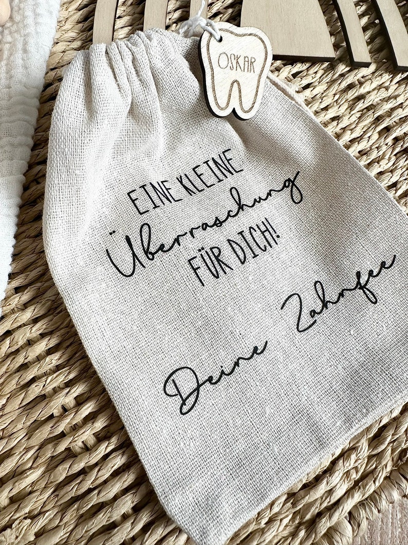 May include: A small white cloth bag with a wooden tooth-shaped tag that says "OSKAR" on it. The bag has black text that says "Eine kleine &Uuml;berraschung f&uuml;r dich! Deine Z&auml;hne".