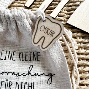 May include: A small white canvas bag with a wooden tooth-shaped tag that says "OSKAR" hanging from it. The bag has the text "EINE KLEINE &Uuml;berraschung F&Uuml;R DICH!" printed on it.