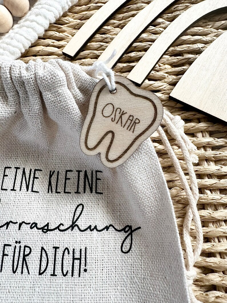 May include: A small white canvas bag with a wooden tooth-shaped tag that says "OSKAR" hanging from it. The bag has the text "EINE KLEINE &Uuml;berraschung F&Uuml;R DICH!" printed on it.