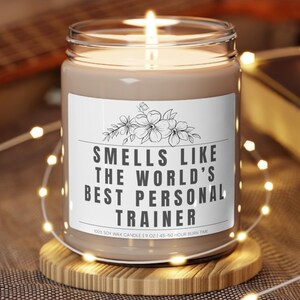 Może przedstawiać: Brązowa świeca sojowa z białą etykietą z napisem "Smells like the world's best personal trainer". Świeca jest zapalona i stoi na drewnianym podkładce.