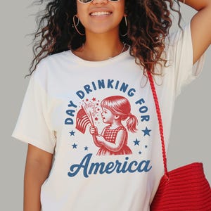 May include: White t-shirt with a red, white, and blue graphic. The graphic features a young girl holding a sparkler and an American flag, with the words "Day Drinking for America" and "America" in blue.