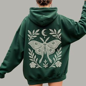 May include: A dark green hooded sweatshirt featuring a large, light-colored graphic on the back. The design displays a central moth surrounded by symmetrical leafy branches, small flowers, and a crescent moon with a star above the moth's head.