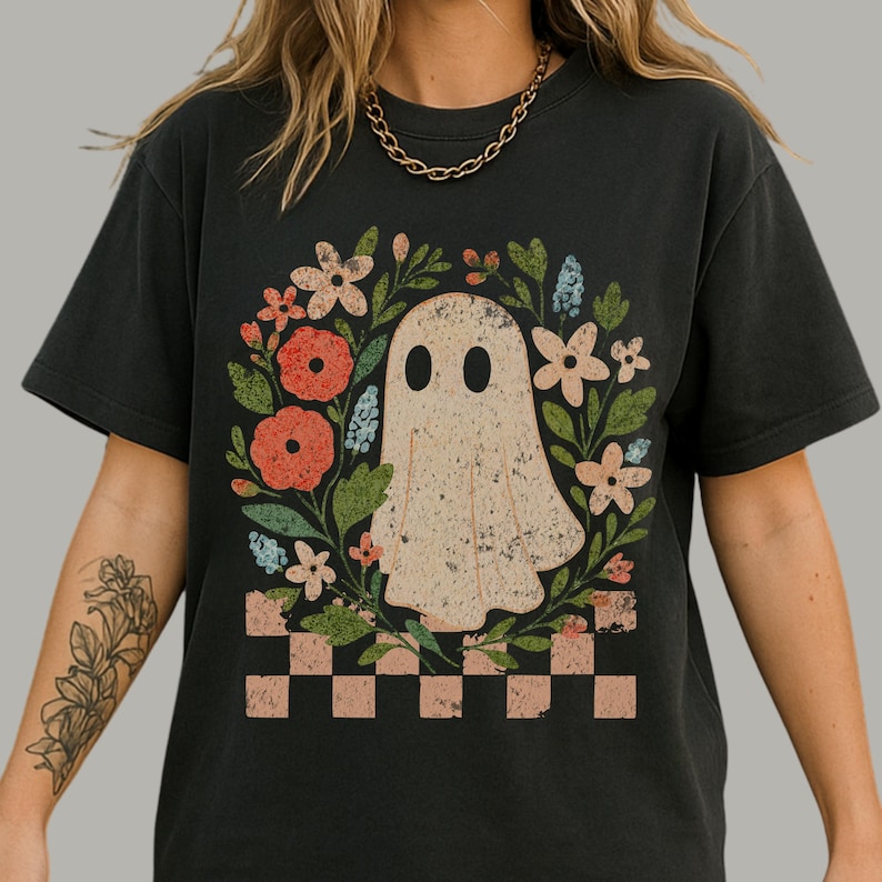 May include: Black t-shirt featuring a ghost illustration surrounded by a floral wreath. The ghost has two black eyes and a vintage, distressed look. The wreath includes red, pink, and blue flowers with green leaves. A checkered pattern is at the bottom.