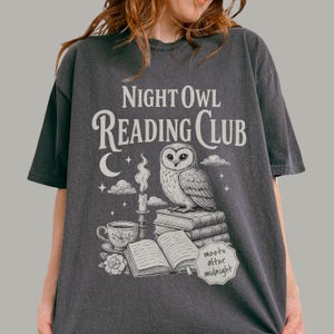 May include: A charcoal gray t-shirt featuring a white graphic print of an owl perched on a stack of books next to a lit candle, a teacup, and a crescent moon. The text reads Night Owl Reading Club and includes a small label saying meets after midnight.