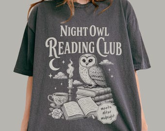 May include: A charcoal gray t-shirt featuring a white graphic print of an owl perched on a stack of books next to a lit candle, a teacup, and a crescent moon. The text reads Night Owl Reading Club and includes a small label saying meets after midnight.
