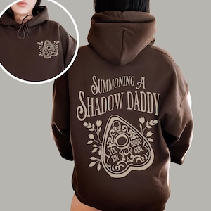 Puede incluir: Una sudadera con capucha marr&oacute;n con cord&oacute;n y bolsillo delantero. La parte trasera de la sudadera presenta el texto "Summoning A Shadow Daddy" sobre un dise&ntilde;o de planchette con las palabras "Yes Sir" y "Good Girl". Un dise&ntilde;o de planchette m&aacute;s peque&ntilde;o est&aacute; en la parte delantera.