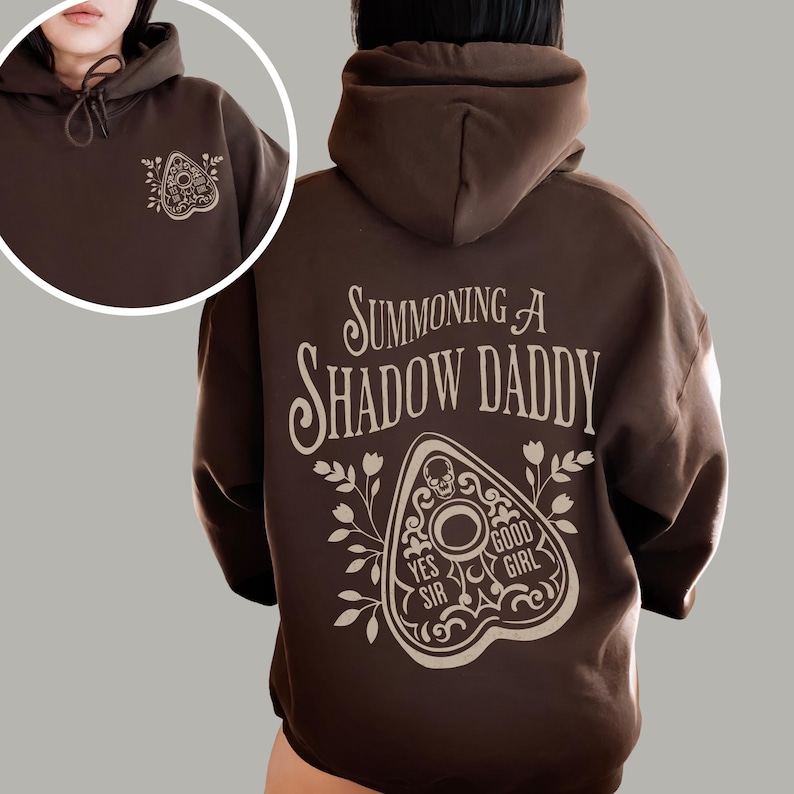 Puede incluir: Una sudadera con capucha marr&oacute;n con cord&oacute;n y bolsillo delantero. La parte trasera de la sudadera presenta el texto "Summoning A Shadow Daddy" sobre un dise&ntilde;o de planchette con las palabras "Yes Sir" y "Good Girl". Un dise&ntilde;o de planchette m&aacute;s peque&ntilde;o est&aacute; en la parte delantera.