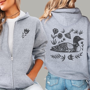 May include: A heather gray zip-up hoodie featuring a small black floral graphic on the front left chest and a large black graphic on the back depicting a loon surrounded by various stylized flowers, leaves, and botanical elements in a block print style.