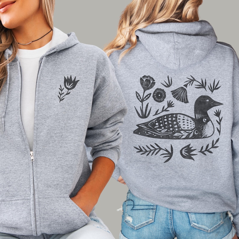 May include: A heather gray zip-up hoodie featuring a small black floral graphic on the front left chest and a large black graphic on the back depicting a loon surrounded by various stylized flowers, leaves, and botanical elements in a block print style.