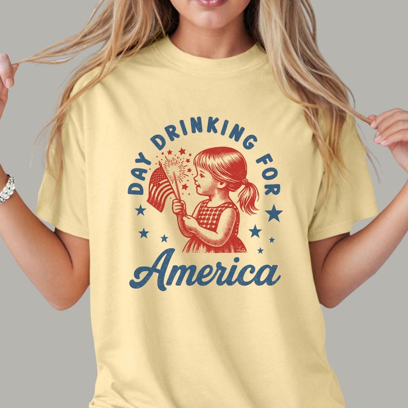 May include: A light yellow t-shirt featuring a vintage-style graphic. The design includes a young girl holding an American flag and fireworks, with the text "Day Drinking for America" in a circular arrangement and the word "America" below.