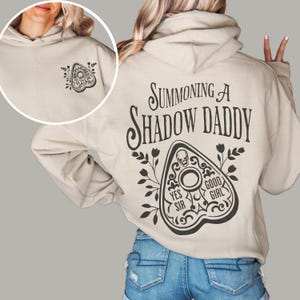 Puede incluir: Sudadera con capucha beige con un gr&aacute;fico negro en la espalda que dice "Summoning A Shadow Daddy". El dise&ntilde;o incluye una planchette con las palabras "Yes Sir" y "Good Girl". Un dise&ntilde;o de planchette m&aacute;s peque&ntilde;o est&aacute; en la parte delantera.