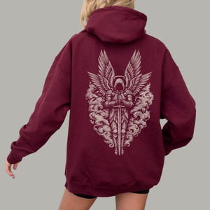 Op de afbeelding: Bordeaux hoodie met een gedetailleerde afbeelding op de rug. Het ontwerp toont een engelachtig figuur met grote vleugels, die een zwaard vasthoudt, omringd door wolkenachtige elementen. De hoodie heeft een capuchon en lange mouwen.