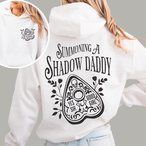 Puede incluir: Sudadera con capucha blanca con un gr&aacute;fico negro en la espalda que dice "Summoning a Shadow Daddy" y un dise&ntilde;o de planchette. La parte delantera de la sudadera tiene un dise&ntilde;o de planchette m&aacute;s peque&ntilde;o. La sudadera tiene capucha y bolsillo delantero.
