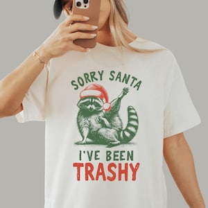 May include: Cream-colored t-shirt featuring a raccoon wearing a Santa hat. The raccoon is illustrated in green and black, with the text "SORRY SANTA I'VE BEEN TRASHY" in green and red. The shirt is a casual, festive design.
