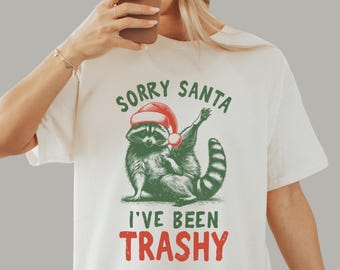 May include: Cream-colored t-shirt featuring a raccoon wearing a Santa hat. The raccoon is illustrated in green and black, with the text "SORRY SANTA I'VE BEEN TRASHY" in green and red. The shirt is a casual, festive design.