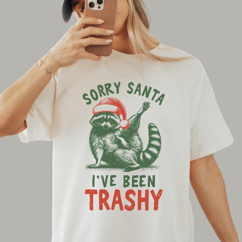 May include: Cream-colored t-shirt featuring a raccoon wearing a Santa hat. The raccoon is illustrated in green and black, with the text "SORRY SANTA I'VE BEEN TRASHY" in green and red. The shirt is a casual, festive design.