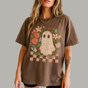 May include: Brown t-shirt featuring a vintage-style ghost illustration surrounded by flowers and a checkered border. The ghost has two oval eyes and a distressed appearance. The shirt is a relaxed fit.