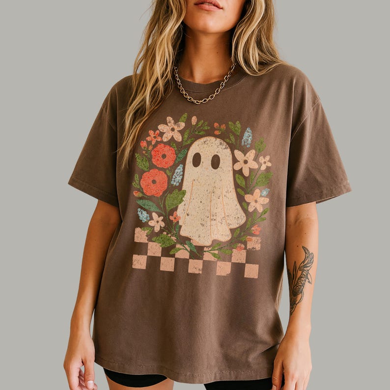 May include: Brown t-shirt featuring a vintage-style ghost illustration surrounded by flowers and a checkered border. The ghost has two oval eyes and a distressed appearance. The shirt is a relaxed fit.