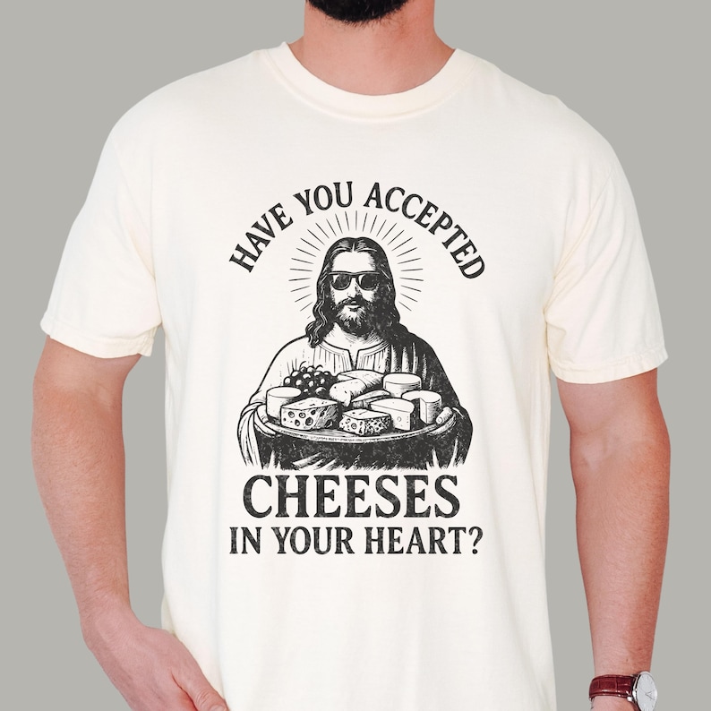 Pu&ograve; includere: Una t-shirt a maniche corte color crema con una grafica nera di una figura barbuta con occhiali da sole che tiene in mano un vassoio di formaggi. Il testo sopra recita HAVE YOU ACCEPTED e il testo sotto recita CHEESES IN YOUR HEART?