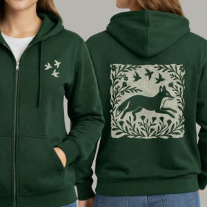 May include: A dark green zip-up hoodie featuring a small white bird graphic on the front chest. The back displays a large square print of a fox running through a field of flowers and foliage, surrounded by three flying birds in a light cream color.