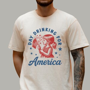 May include: Cream-colored t-shirt featuring a vintage-style graphic. The design includes a young girl holding a sparkler and an American flag, with the words "Day Drinking for America" in a circular arrangement.