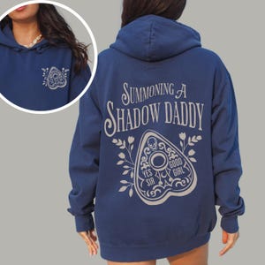 Puede incluir: Sudadera con capucha azul marino con el texto "Summoning a Shadow Daddy" en la espalda. Un dise&ntilde;o de planchette blanco est&aacute; debajo del texto. La parte delantera de la sudadera tiene un dise&ntilde;o de planchette m&aacute;s peque&ntilde;o.