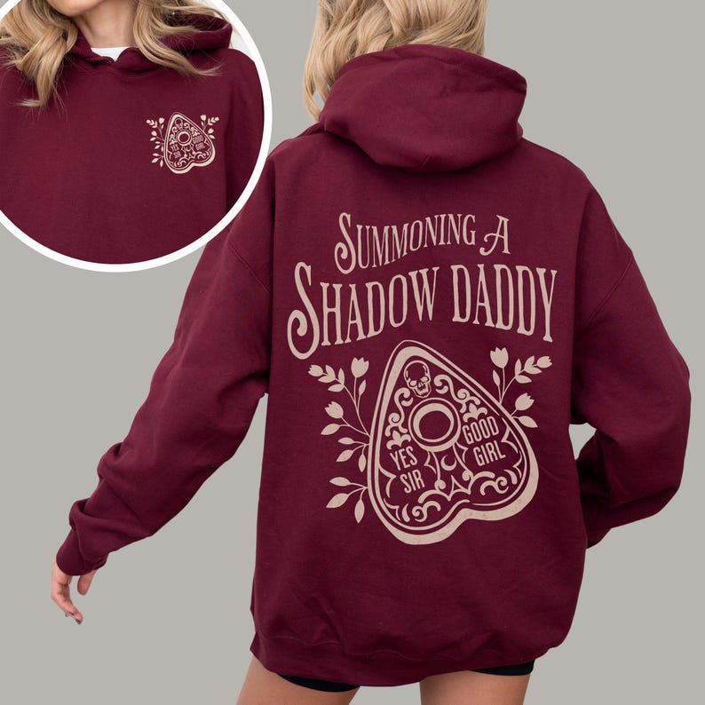 Puede incluir: Sudadera con capucha burdeos con el texto "Summoning a Shadow Daddy" en la espalda, junto con un dise&ntilde;o de planchette. Un dise&ntilde;o de planchette m&aacute;s peque&ntilde;o est&aacute; en la parte delantera. La sudadera con capucha es de color s&oacute;lido y tiene una capucha.