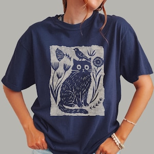 May include: A navy blue short-sleeve t-shirt featuring a central rectangular graphic print. The design displays a dark blue illustration of a cat sitting with a small bird perched on its head, surrounded by stylized floral and botanical elements.
