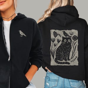 May include: A black zip-up hoodie featuring a small bird graphic on the left chest. The back displays a large, rectangular block print illustration of a cat sitting among stylized flowers with a bird perched on its head, all in black ink.
