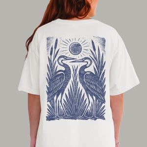 May include: An off-white short-sleeve t-shirt featuring a large blue graphic print on the back. The design depicts two long-necked birds facing each other amidst tall reeds and cattails, with a stylized sun positioned above the birds.