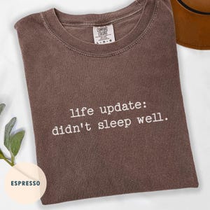 May include: Espresso-colored t-shirt with the text "life update: didn't sleep well" in white. The shirt has a crew neck and a Comfort Colors tag.