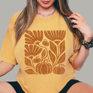 May include: A mustard yellow short-sleeve t-shirt featuring a large, centered graphic print in a distressed brown ink. The design displays a collection of stylized flowers, leaves, and pumpkins arranged in a square composition on the chest.