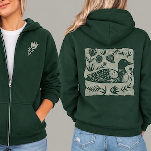 May include: A dark green zip-up hoodie featuring a small light green floral graphic on the left chest and a large square print on the back depicting a stylized loon bird surrounded by various botanical elements in a light green color.