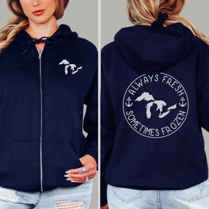 May include: Navy blue zip-up hoodie with a white embroidered outline of the Great Lakes on the front. The back features a circular design with the text "ALWAYS FRESH SOMETIMES FROZEN" around a Great Lakes outline, with anchors on each side. The hoodie has a drawstring hood.