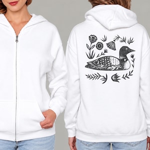May include: A white zip-up hoodie featuring a black graphic print on the back. The design depicts a loon bird surrounded by stylized floral and leaf elements. The front of the hoodie includes a full-length silver zipper and two front pockets.