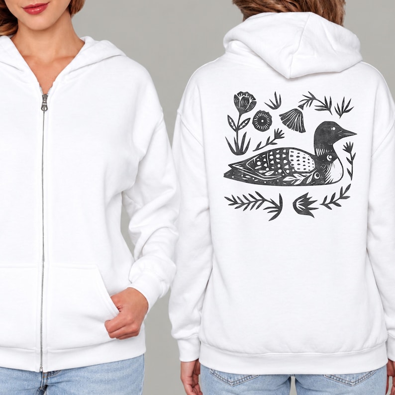 May include: A white zip-up hoodie featuring a black graphic print on the back. The design depicts a loon bird surrounded by stylized floral and leaf elements. The front of the hoodie includes a full-length silver zipper and two front pockets.