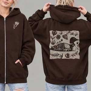 May include: A dark brown zip-up hoodie featuring a small light-colored flower graphic on the front left chest. The back displays a large, rectangular, light-colored patch with a detailed black illustration of a loon surrounded by various floral motifs.