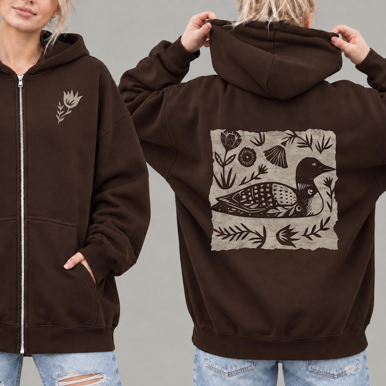 May include: A dark brown zip-up hoodie featuring a small light-colored flower graphic on the front left chest. The back displays a large, rectangular, light-colored patch with a detailed black illustration of a loon surrounded by various floral motifs.